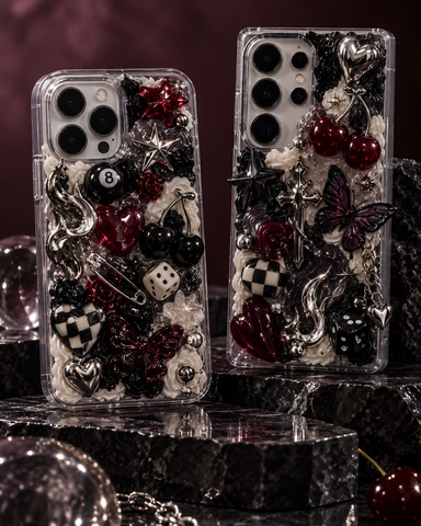 Two clear smartphone cases filled with gothic-themed embellishments, including black and red cherries, silver and black dice, hearts, stars, checkered pieces, and butterflies, displayed on a dark marble surface against a dark burgundy background.